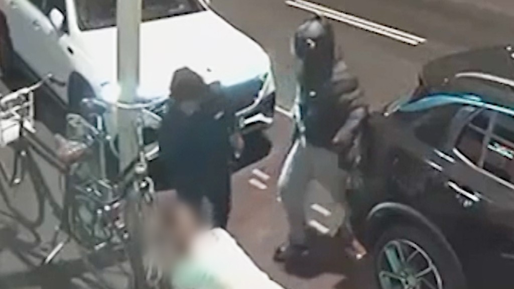 twee personen, donker gekleed, staan tussen auto's bij een voetpad